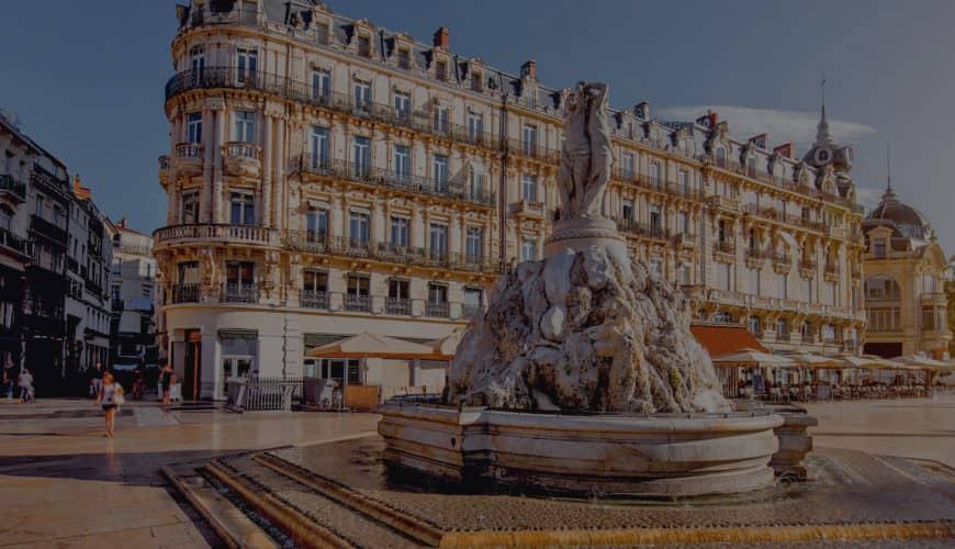 Place avec fontaine centrale, bâtiment historique, passants et terrasses de café visibles.