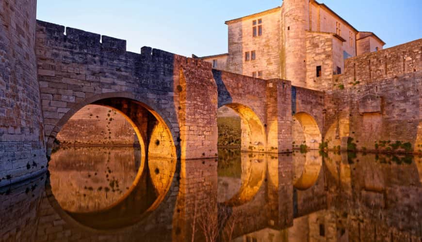 Pont en pierre avec arches et bâtiment historique illuminé, reflet dans l'eau calme.