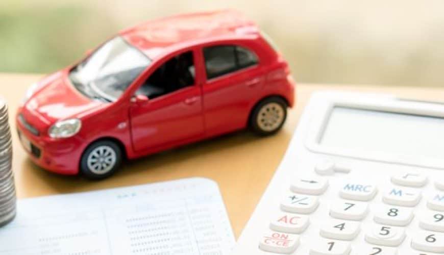 Calculatrice, modèle réduit de voiture rouge, pièces empilées et carnet sur une table en bois.