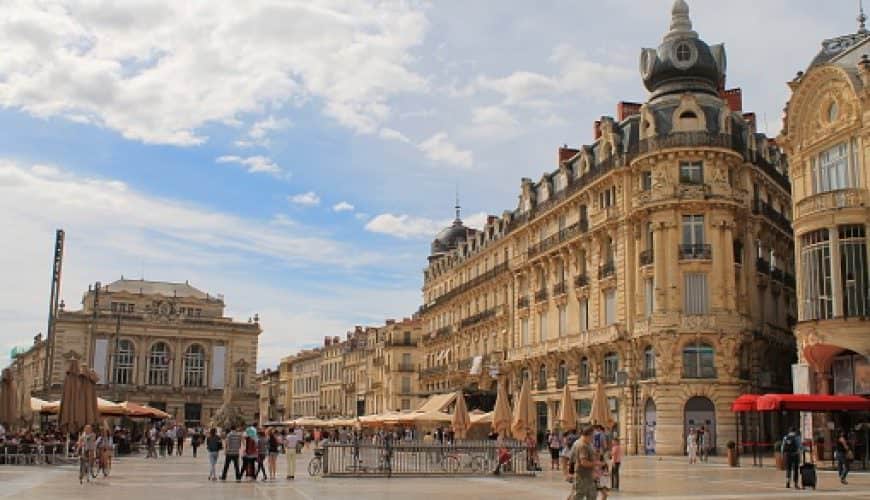 Grande place urbaine avec bâtiments historiques, terrasses de café, passants, ciel nuageux.