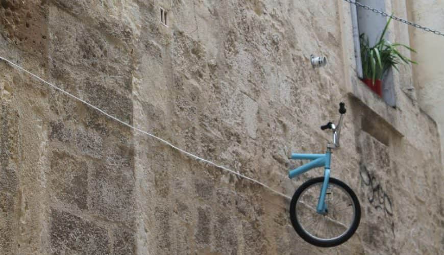 Turquoise bicycle wheel hanging on a stone wall near an electrical wire and window with potted plant.