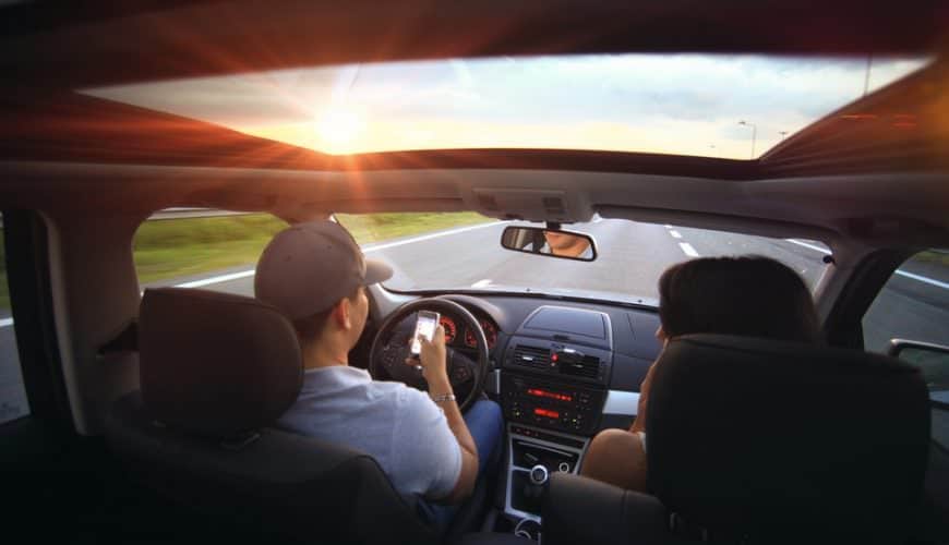 Conducteur et passagère dans une voiture, conducteur tenant un téléphone sur route ensoleillée.