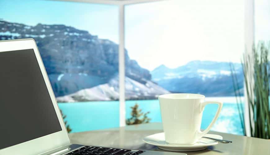 Ordinateur portable et tasse sur table, vue sur montagnes enneigées et eau turquoise.