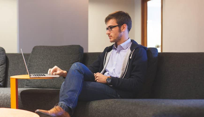 Homme avec lunettes sur canapé sombre, utilise un portable sur table basse orange, intérieur moderne.