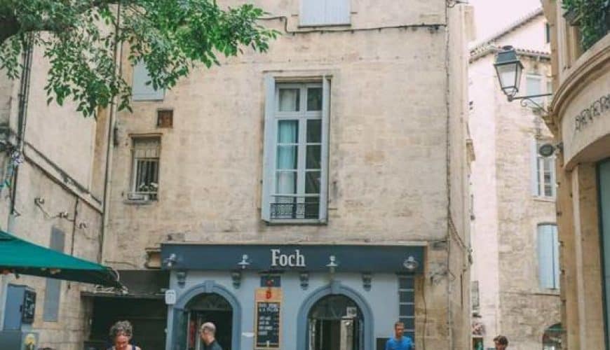 People sitting at Foch café terrace in a sunny, narrow street with stone buildings and a tree.