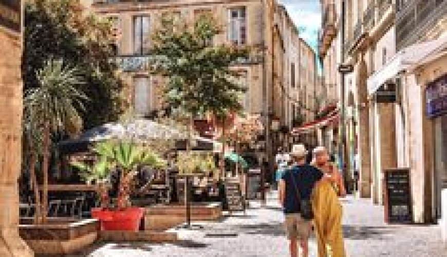 Rue pittoresque avec bâtiments en pierre, terrasses de café, plantes et passants au soleil.