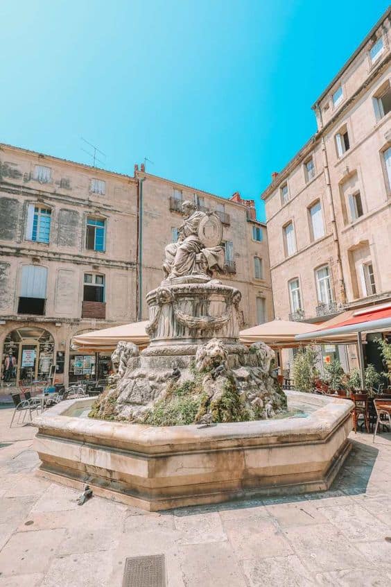 Stone fountain with sculptures in a plaza, surrounded by old buildings and café tables.