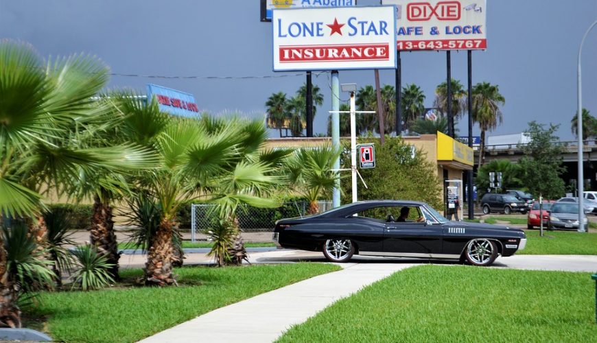 Voiture classique noire près de palmiers et panneaux publicitaires locaux en arrière-plan.