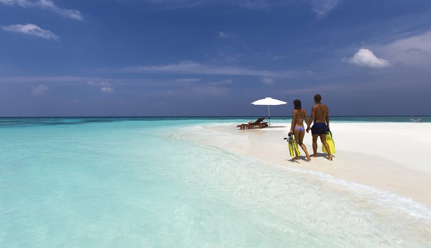 Personnes marchant sur plage sable blanc avec palmes, eau turquoise, parasol et chaises longues.