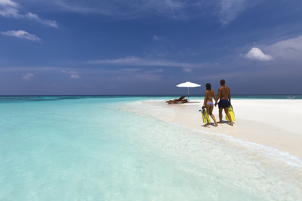 Personnes marchant sur plage sable blanc avec palmes, eau turquoise, parasol et chaises longues.