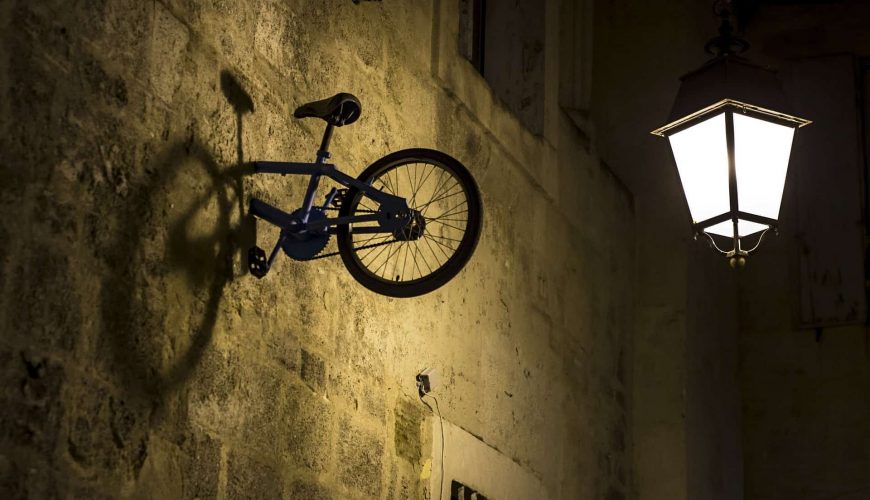 Bicyclette fixée verticalement sur un mur en pierre avec un lampadaire éclairé à côté.