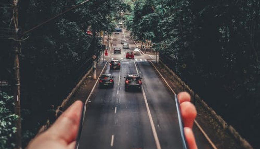 Main tenant un téléphone avec écran reflétant une route bordée d'arbres et des voitures.