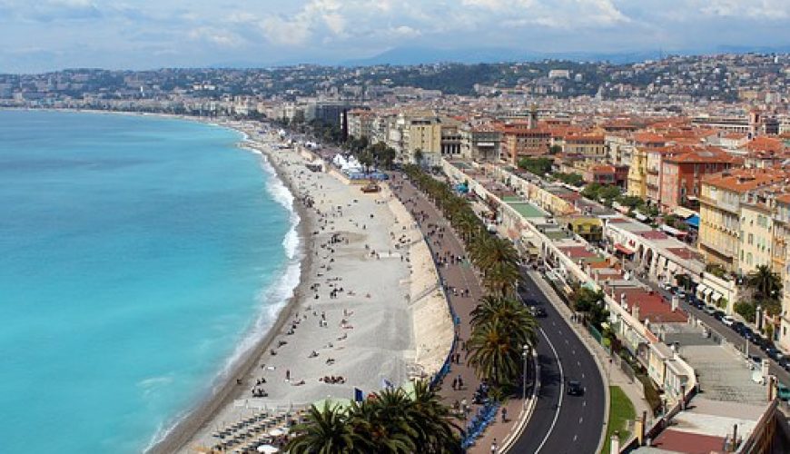 Vue aérienne d'une ville côtière avec promenade, palmiers, plage de sable et mer turquoise.