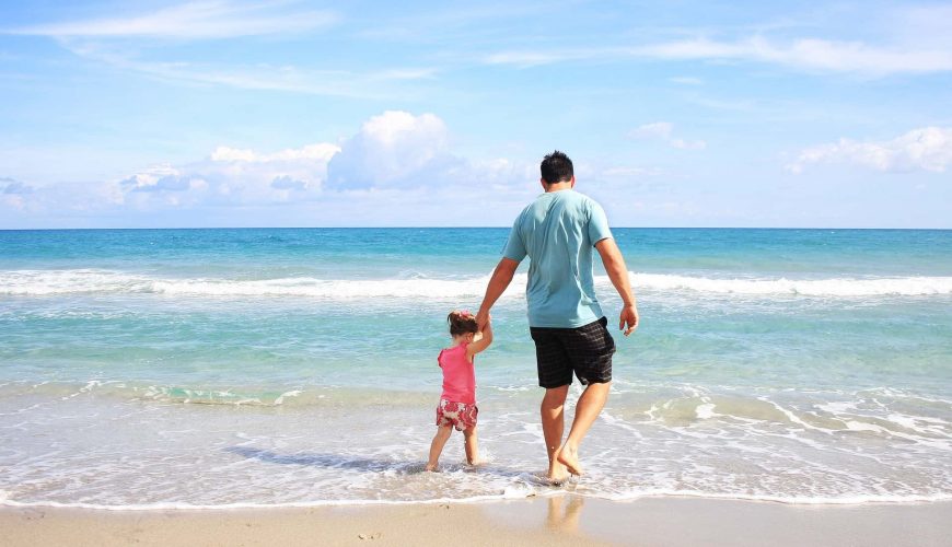 Adulte et enfant marchant main dans la main sur une plage ensoleillée avec l'océan derrière.