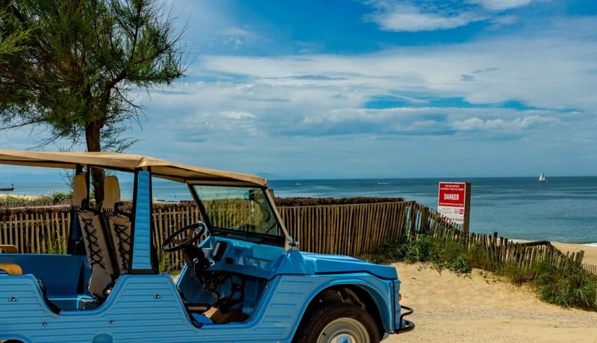 Voiture décapotable bleue près de la plage avec ciel partiellement nuageux en arrière-plan.