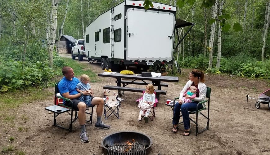 Famille autour d'un feu de camp en forêt avec un camping-car et une table de pique-nique.