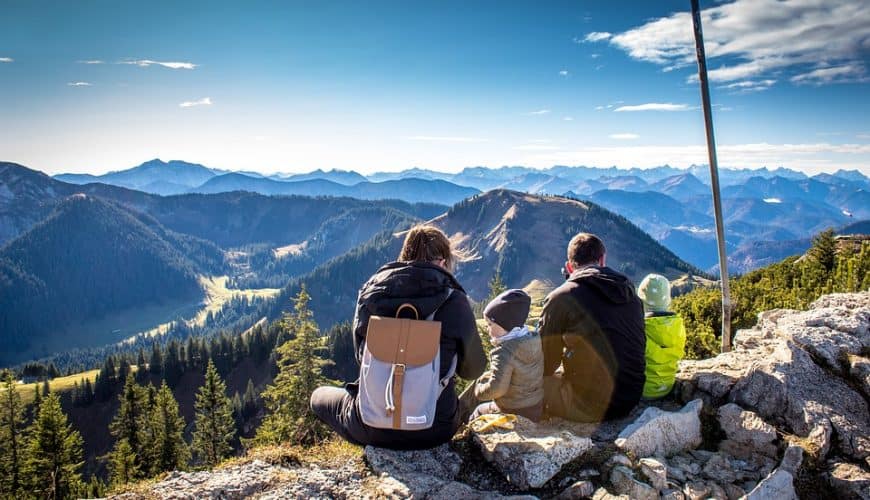 Famille assise sur un sommet montagneux admirant un paysage avec ciel bleu et montagnes.