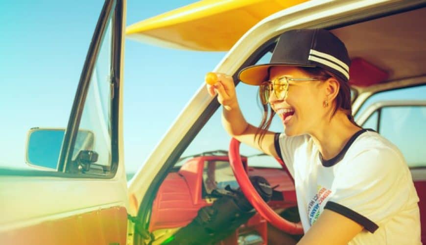 Personne souriante dans voiture ancienne, casquette, lunettes de soleil, tenant une orange.