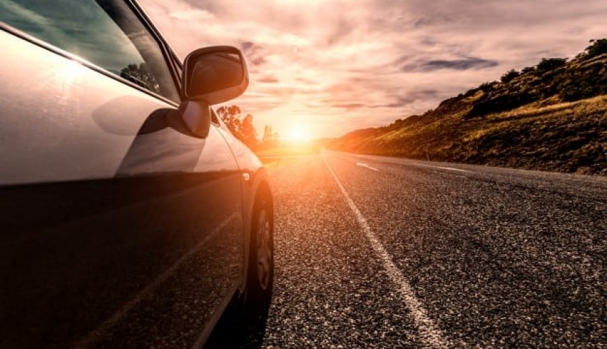 Voiture sur route sous coucher de soleil, ciel partiellement nuageux, collines à l'horizon.