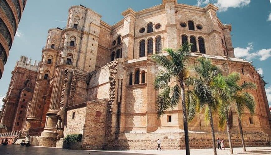 Imposing stone building with ornate details, palm trees, under a partly cloudy blue sky.