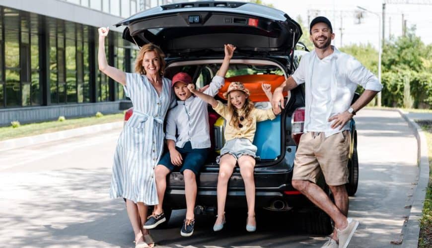 Famille joyeuse avec deux enfants posant près d'une voiture ouverte sur une route ensoleillée.