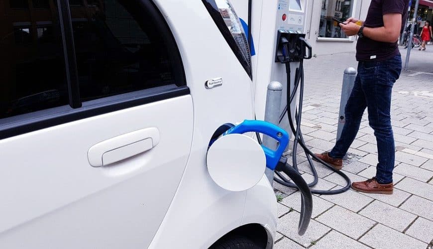 Voiture blanche en charge à une borne électrique, câble bleu, personne avec téléphone.