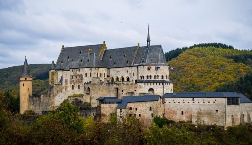 Château médiéval entouré de collines verdoyantes avec drapeaux visibles, sous un ciel nuageux.