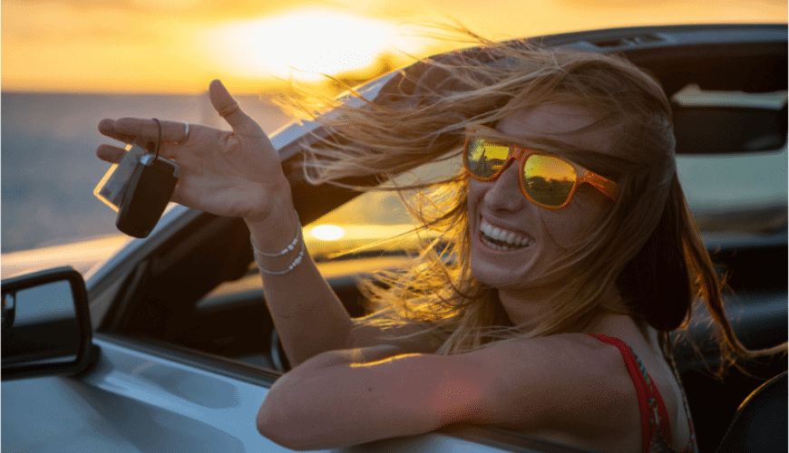 Femme souriante avec lunettes de soleil dans une décapotable, tenant des clés au coucher du soleil.