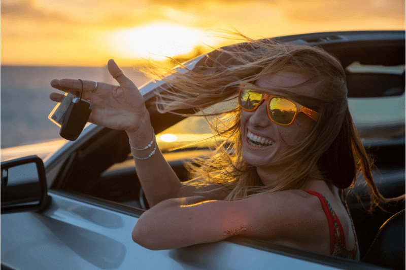 Femme souriante avec lunettes de soleil dans une décapotable, tenant des clés au coucher du soleil.