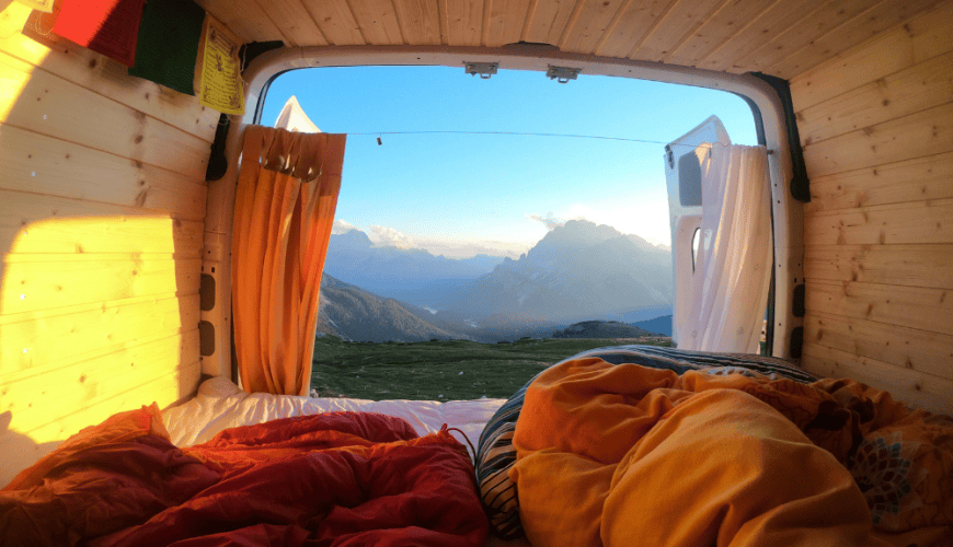 Intérieur d'un van en bois avec lit coloré, rideaux, vue sur montagnes et ciel dégagé.