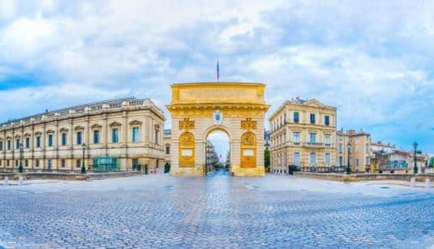 Place pavée avec arc de triomphe central, entourée de bâtiments historiques, ciel partiellement nuageux.