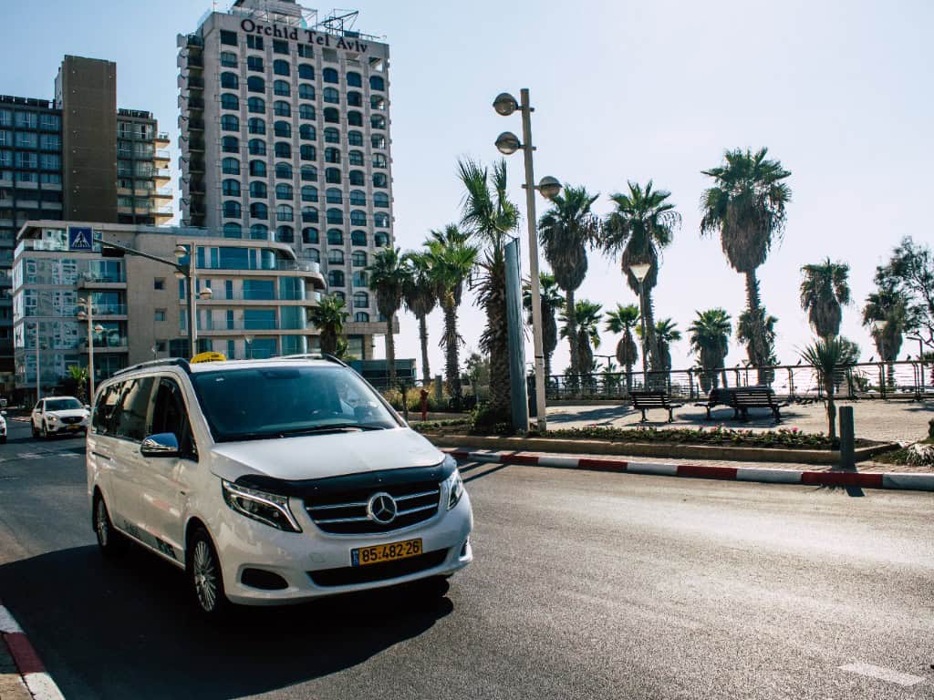 Voiture blanche Mercedes plaque jaune sur route avec palmiers, hôtel Orchid Tel Aviv adjacent.