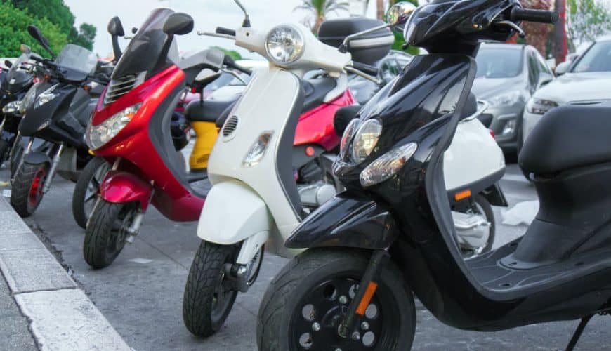 Range of colorful scooters parked on an urban sidewalk, among cars and greenery